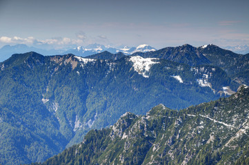 Pine forests, rocks and snow patches on the steep slopes of Karavanke / Karawanken mountain range in Slovenia, with Carnic Alps of Austria and Italy in the background