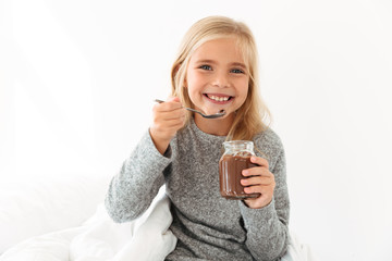 Happy blonde girl in gray pajamas holding glass jar and spoon with chocolate spread, looking at camera