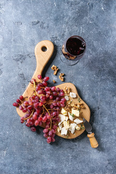 Wooden serving board with fresh red grapes, walnuts, goat and cheddar cheese, with cheese knife and glass of red wine over blue texture background. Top view, copy space.