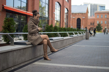 Fototapeta na wymiar Young woman in a beige coat is sitting on a bench, smiling and looking at the phone.