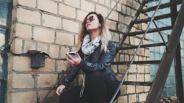 Smiling woman in sunglasses, a black leather jacket, black jeans standing on an urban metal stair against a brick wall and dances. Woman listening to music on the stairs of industrial building.