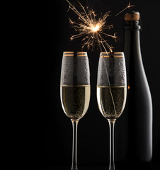 A bottle of champagne and two filled glasses on a black background, in one glass a Bengal fire burns.