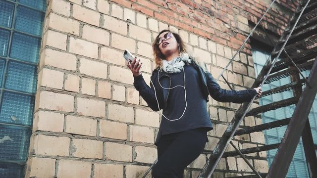 Smiling woman in sunglasses, a black leather jacket, black jeans standing on an urban metal stair against a brick wall and dances. Woman listening to music on the stairs of industrial building.