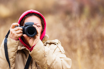 Fototapeta premium Female photographer looking through the viewfinder of the camera. The woman is dressed in a jacket and hood. On the street the cold season