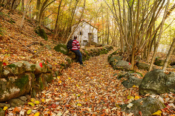 Fototapeta premium Piedicavallo, Italy - October 20, 2017: Young man observes the old rustic house along an alpine trail full of autumn colors
