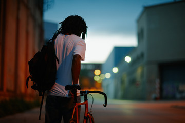 Young afro man walking with a bike in the street at dawn