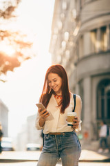 Young woman with coffee to go standing at the street and using mobile phone  