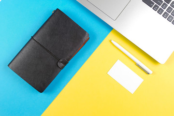 Top view of laptop computer, paper travel notebook with blank card and white pen on blue and yellow color background