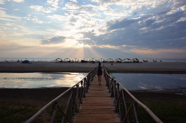 Obraz premium dziewczyna odchodzi w dal do oceanu zachodzącego słońca i plaży wzdłuż długiego drewnianego mostu