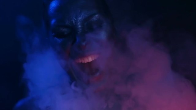 Halloween night. Horror Vape Party. Beautiful Sexy Young vamp Woman with glamorous mystical makeup vaping in Nightclub (exhaling smoke). Girl smoking in Club. Blue mystic smoke