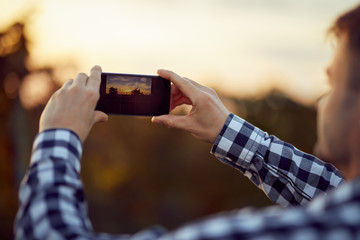 Back view of man taking photo with digital camera on mobile phone of sunset