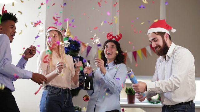 Happy multi-ethnic people in funny hats lighting sparklers celebrating christmas and new year in the office, excited diverse young friends holding champagne glasses, firing bengal lights