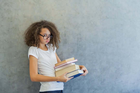 Curly hair teen girl with glasses stands next to the wall and holds several books