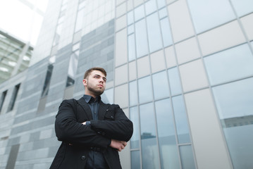 Business portrait of a young man in a suit from the lower angle against the backdrop of modern architecture. A beautiful man with a beard poses against the background of the city.