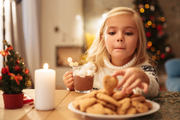 Funny little girl with cup of hot chocolate taking biscuit from plate while sitting at festive table