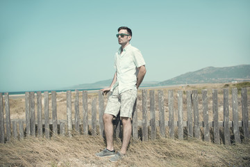 Retrato de hombre joven con gafas de sol vestido en colores claros al aire libre junto a una playa