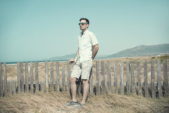 Retrato de hombre joven con gafas de sol vestido en colores claros al aire libre junto a una playa