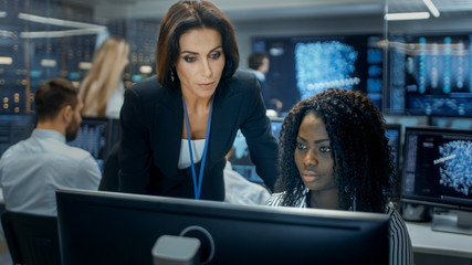Female Team Leader Consults Young Computer Engineer. They Work in a Crowded Office on a Neural Network/ Artificial Intelligence Project.