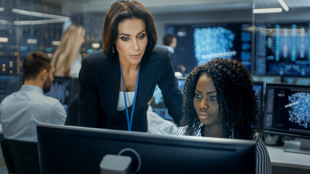 Female Team Leader Consults Young Computer Engineer. They Work in a Crowded Office on a Neural Network/ Artificial Intelligence Project.