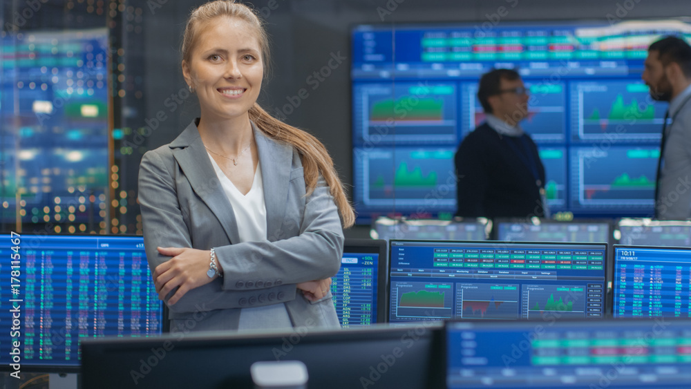 Poster successful female stock trader crosses arms and smiles at the camera. in the background busy stock e