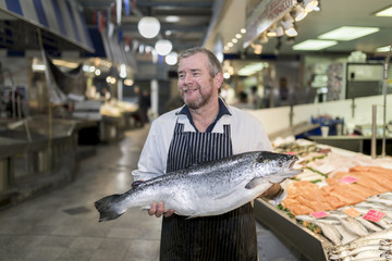 Male fishmonger wearing an apron holding large and whole salmon fish in front of display counter...