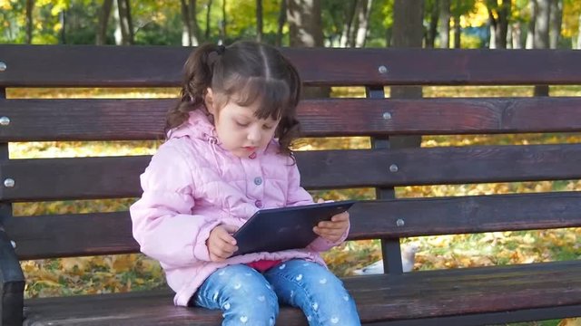 A child with a tablet in the park. Little girl on a bench with a tablet on nature.