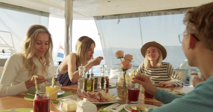 Group of Young Beautiful People Eating Healthy Breakfast on a Yacht, They're Friendly Chatting. They Have Various Dishes and Smoothies Served on the Table. Sea is in the Background. 