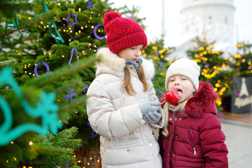 Two adorable little sisters eating red apples covered with sugar icing on traditional Christmas market