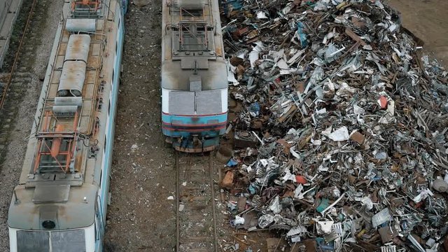 large heaps of metal scrap and containers with garbage are near a railway with trains in day in cloudy weather,