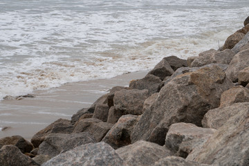 Large rocks line the seaside ocean coast beach as waves crash up against from the current