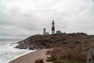 Wide establishing view of beautiful ocean coast lighthouse standing tall over rocky beach. Tourists and people fish and walk the rocky shoreline during the day
