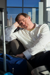 Tired Caucasian businessman sleeping at airport terminal next to luggage, sunlight streaming in through glass windows