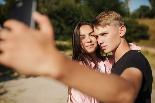 Portrait of young beautiful couple standing and making selfie in park. Cute couple taking photos on cellphone frontal camera