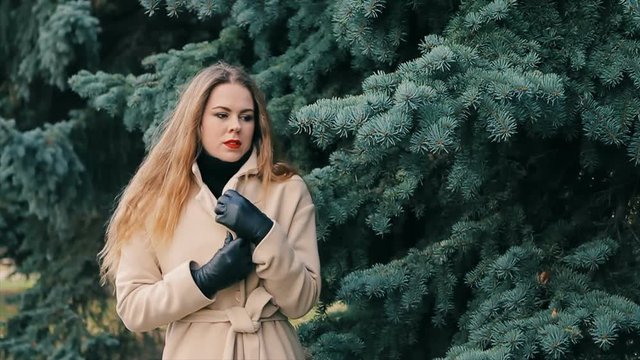 Beautiful Young blonde woman looking like Nicole Kidman in winter park with pine slow motion. Girl in cream coat and black leather gloves walking near green and blue pine, posing for camera.