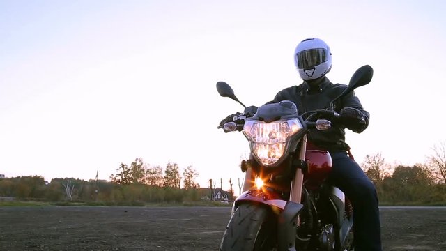 A young man in black leather jacket and white helmet sits on motorcycle turns on lights before journey at autumn sunset. Steady cam shot.