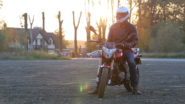 A young man in black leather jacket and white helmet sits on motorcycle turns on lights before journey at autumn sunset.