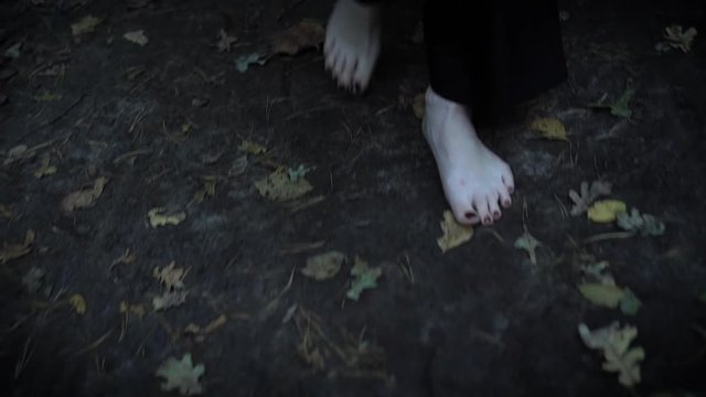 bare pale feet of woman is wearing long black coat, walking in forest in autumn over wet earth and fallen leaves