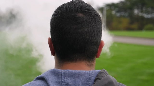 A rear view of a young millennial man using his e-cigarette (vape pen) outside on an overcast day. Shot in slow motion.  	