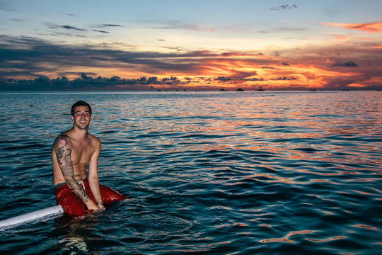 Young man with swimsuit and tattoed arm sitting on the edge of a catamaran boat on the sea at sunset