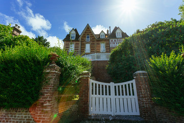 Country house with green fence and white gate in Normandy, France on a sunny day with sunbeam in blue sky. Beautiful countryside, lifestyle and typical french architecture, european country landscape.