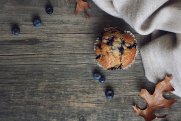 Still life with blueberry muffin, warm blanket, leaves and blueberries over rustic wooden background shot from directly above, copy space, horizontal
