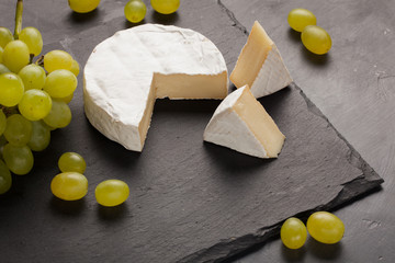 Cutting board of camembert cheese and white grapes on dark stone background