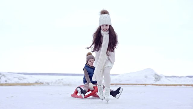 Adorable girl in pink coat and warm hat pulling little brother on sled while skating on ice on outdoor rink in winter