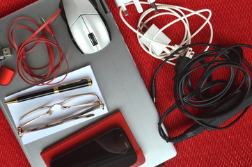 Technology for travel. Closed laptop computer with chargers, mouse, glasses, pen, notebook, phone and USB stick on red background.