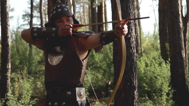 Archer with an eastern appearance aiming an arrow in forest. Mongolian warrior with leather armor pulls string on a small bow and lets it go.