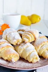 Homemade croissants with fruit jam, decorated with powdered sugar on a ceramic plate and cup of morning tea on a white wooden background.