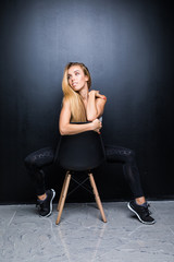 Fototapeta premium Sexy and beautiful young blonde model with slim body is posing in the studio on the chair, dark wall on the background
