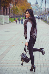 Happy young adult woman smiling outdoors and walking on city street at sunset time wearing winter clothes and fur jacket, winter holidays and fashionable people concept, winter time at city