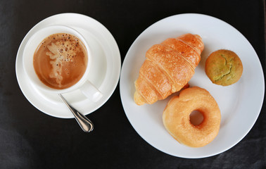 Cup of hot coffee with Croissant, donut and muffin in white plate.