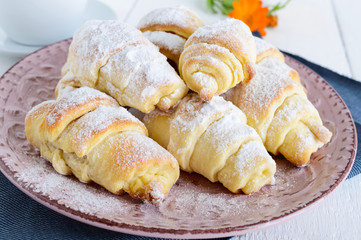 Homemade croissants with fruit jam, decorated with powdered sugar on a ceramic plate and cup of morning tea on a white wooden background.